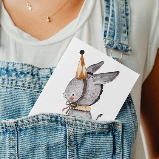 Person in weißem Shirt mit blauer Latzhose hält Karte Esel Vintage A6 mit Esel im Partyhut; ideal als Gruß- oder Geschenkkarte.