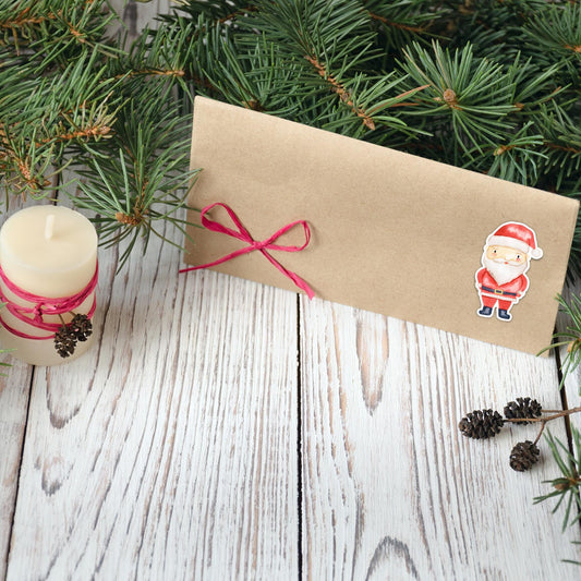 Sticker Weihnachtsmann auf einem kraftpapier Umschlag mit rotem Band, umgeben von Tannenzweigen, Kerze und Tannenzapfen – ideal für Weihnachtskarten und Dekoration.