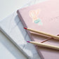 Sticker Meerjungfrau rosa auf einem pinken Notizbuch mit zwei Bleistiften, zeigt handgemaltes Meerjungfrauenmotiv in Pastellfarben, ideal für kreative Verschönerungen.