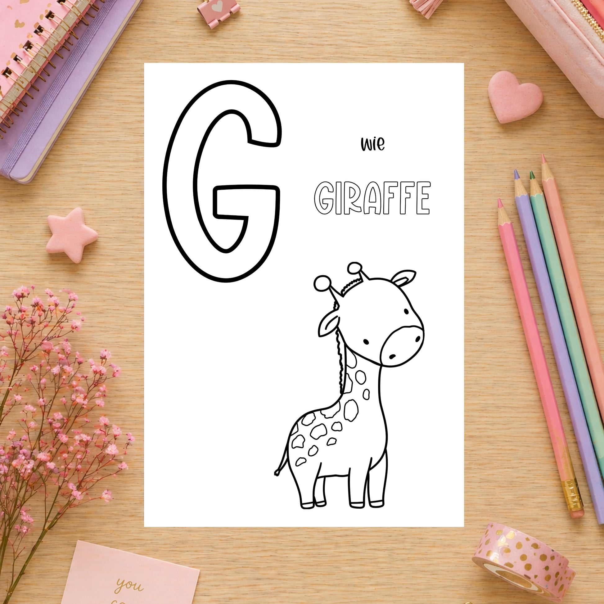 Tierisches Alphabet – Ausmalbilder zum Ausdrucken: Flachlage mit Buchstaben G, Giraffe-Ausmalbild, pastellfarbene Notizbücher, Bleistifte, Radiergummis, Blumen und Zubehör auf Holztisch.