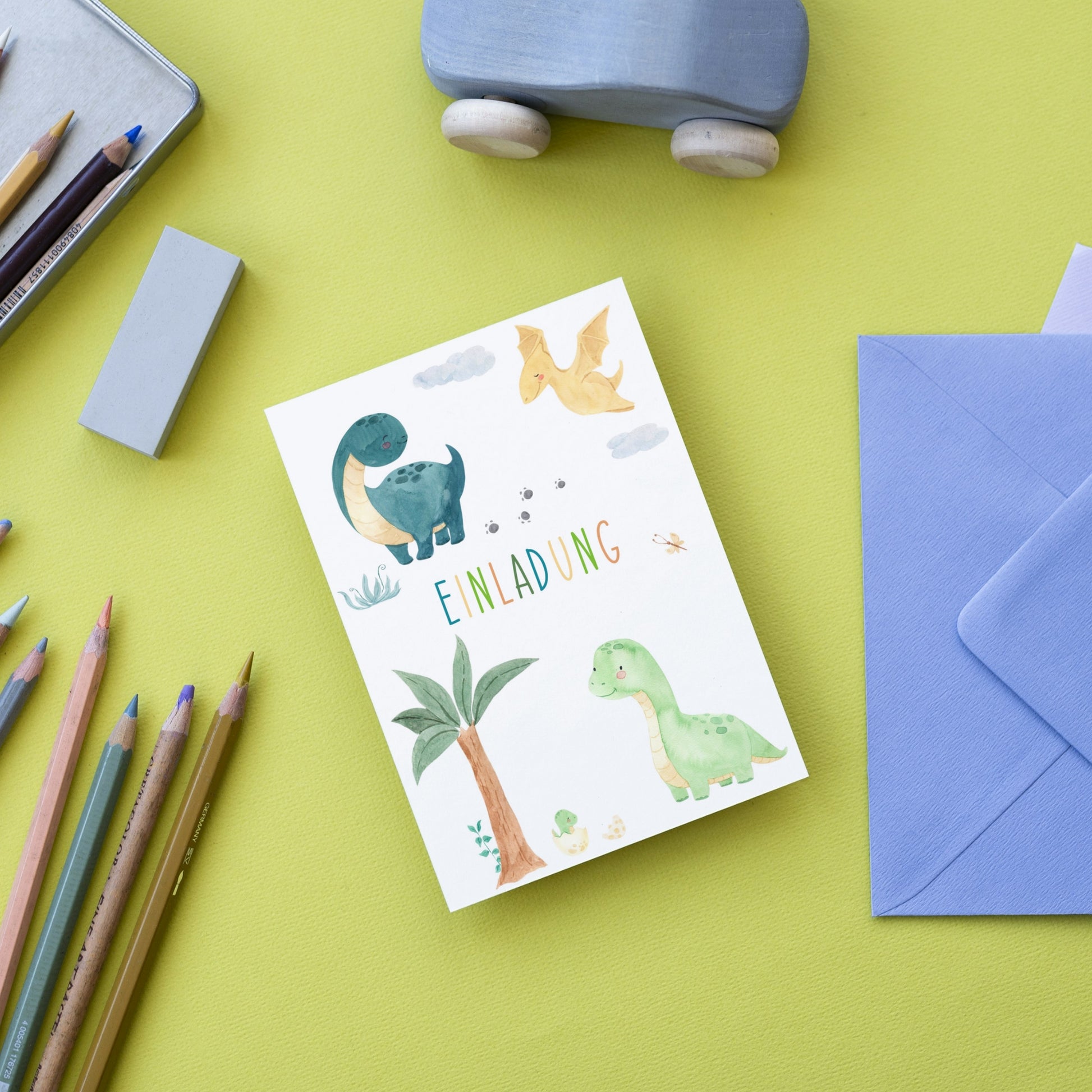 Einladung Kindergeburtstag Dinosaurier A6: Karte mit Cartoon-Dinosauriern, blauer Umschlag, Bleistifte und Spielzeugauto; hochwertige 300g Naturpapierkarte für Kindergeburtstage.