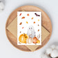 Handgemalte Halloweenkarte mit Aquarell-Gespenst, Kürbissen, Herbstlaub und Fledermaus auf braunem Umschlag auf Holzbrett.