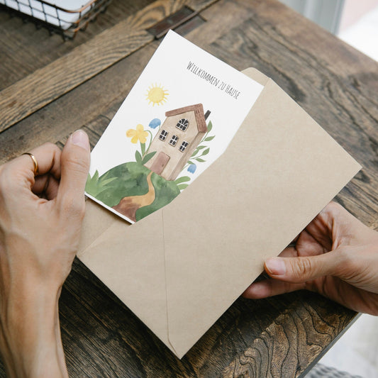 Eine Person hält Postkarte mit Aquarellmotiv: Hause auf einem Hügel, gedruckt auf hochwertigem, mattem 300g Naturpapier, Größe A6, beschreibbar und bestempelbar, ideal für Einzug/Umzug.