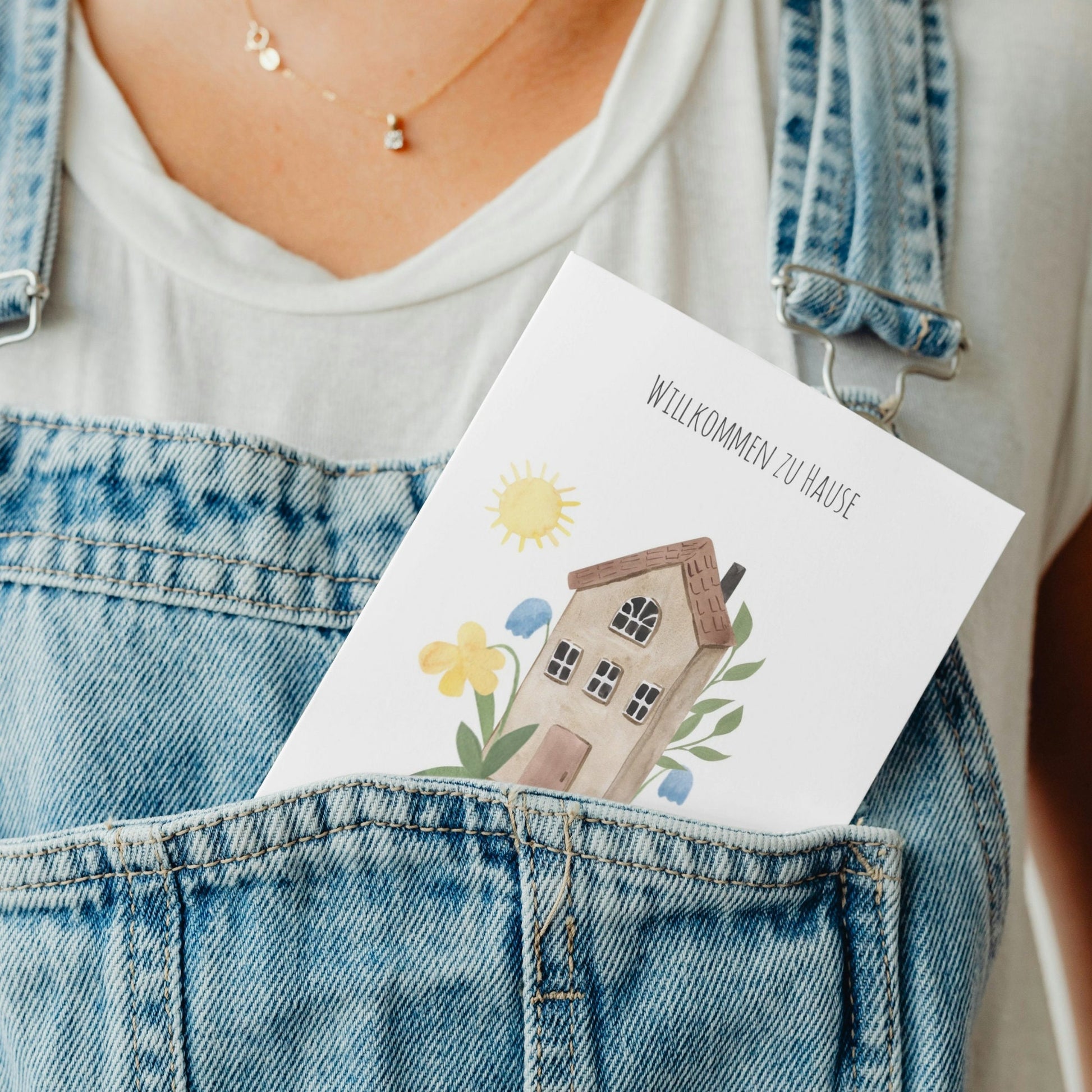 Person trägt weiße Bluse und blaue Latzhose. In der Tasche Postkarte mit Aquarellmotiv: Hause auf einem Hügel, gedruckt auf hochwertigem, mattem 300g Naturpapier, Größe A6, beschreibbar und bestempelbar, ideal für Einzug/Umzug.