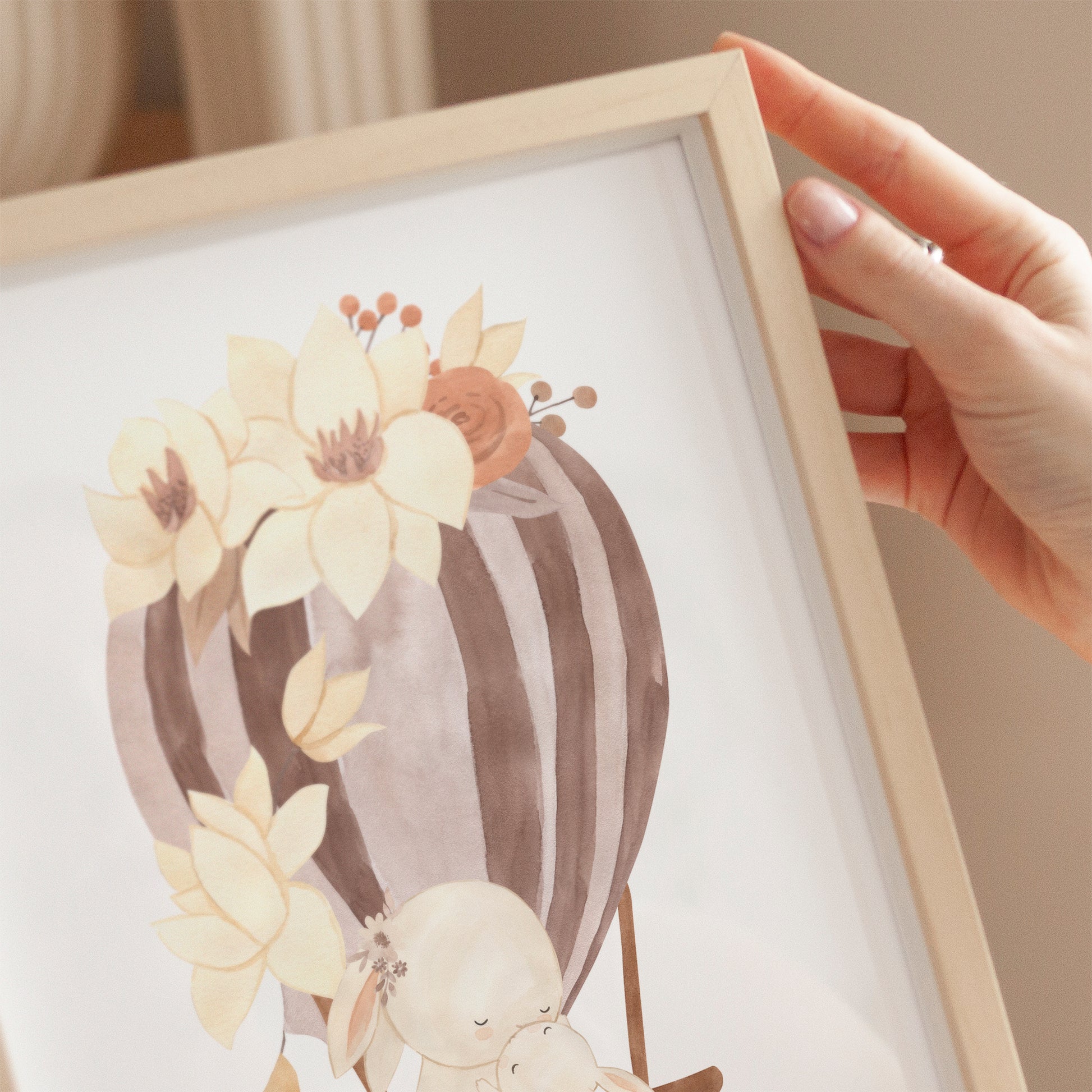 Person hält Poster Heißluftballon Mama und Baby Hase A4 & A3: Aquarellbild von Mama Hase und Baby in blumenverziertem Heißluftballon, ideal als Kinderzimmerdeko auf hochwertigem Naturpapier.