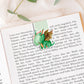 Magnetisches Lesezeichen Drache: Handgemaltes Drachenmotiv, ca. 6 x 4 cm, auf 200g Papier gedruckt, mit Vinyl versiegelt, enthält Magnete zum sicheren Halt. Auf Buchseite präsentiert.