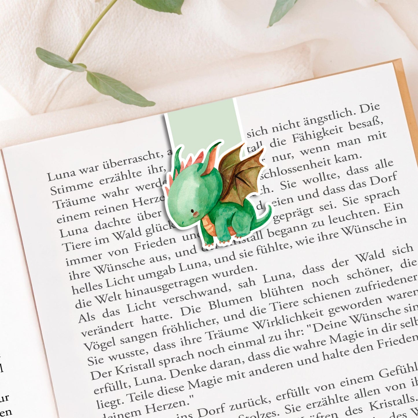 Magnetisches Lesezeichen Drache: Handgemaltes Drachenmotiv, ca. 6 x 4 cm, auf 200g Papier gedruckt, mit Vinyl versiegelt, enthält Magnete zum sicheren Halt. Auf Buchseite präsentiert.