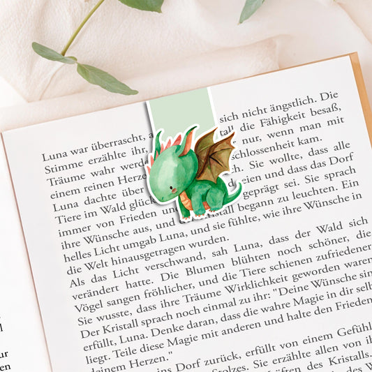 Magnetisches Lesezeichen Drache: Handgemaltes Drachenmotiv, ca. 6 x 4 cm, auf 200g Papier gedruckt, mit Vinyl versiegelt, enthält Magnete zum sicheren Halt. Auf Buchseite präsentiert.