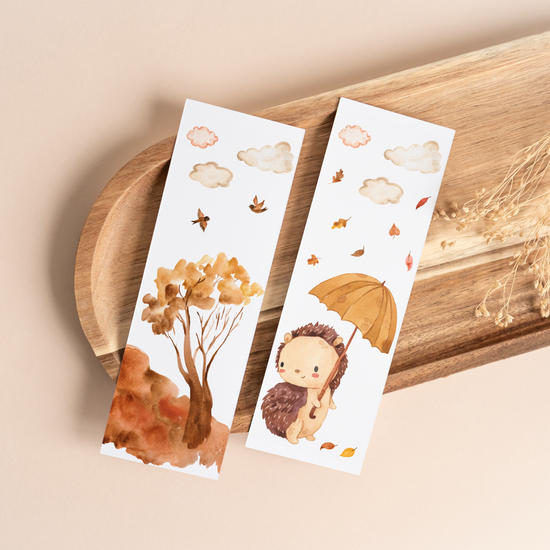Zwei herbstliche Lesezeichen mit Wasserfarben-Illustrationen: ein Herbstbaum mit Vögeln und ein Igel unter einem Regenschirm. Auf Holztablett.