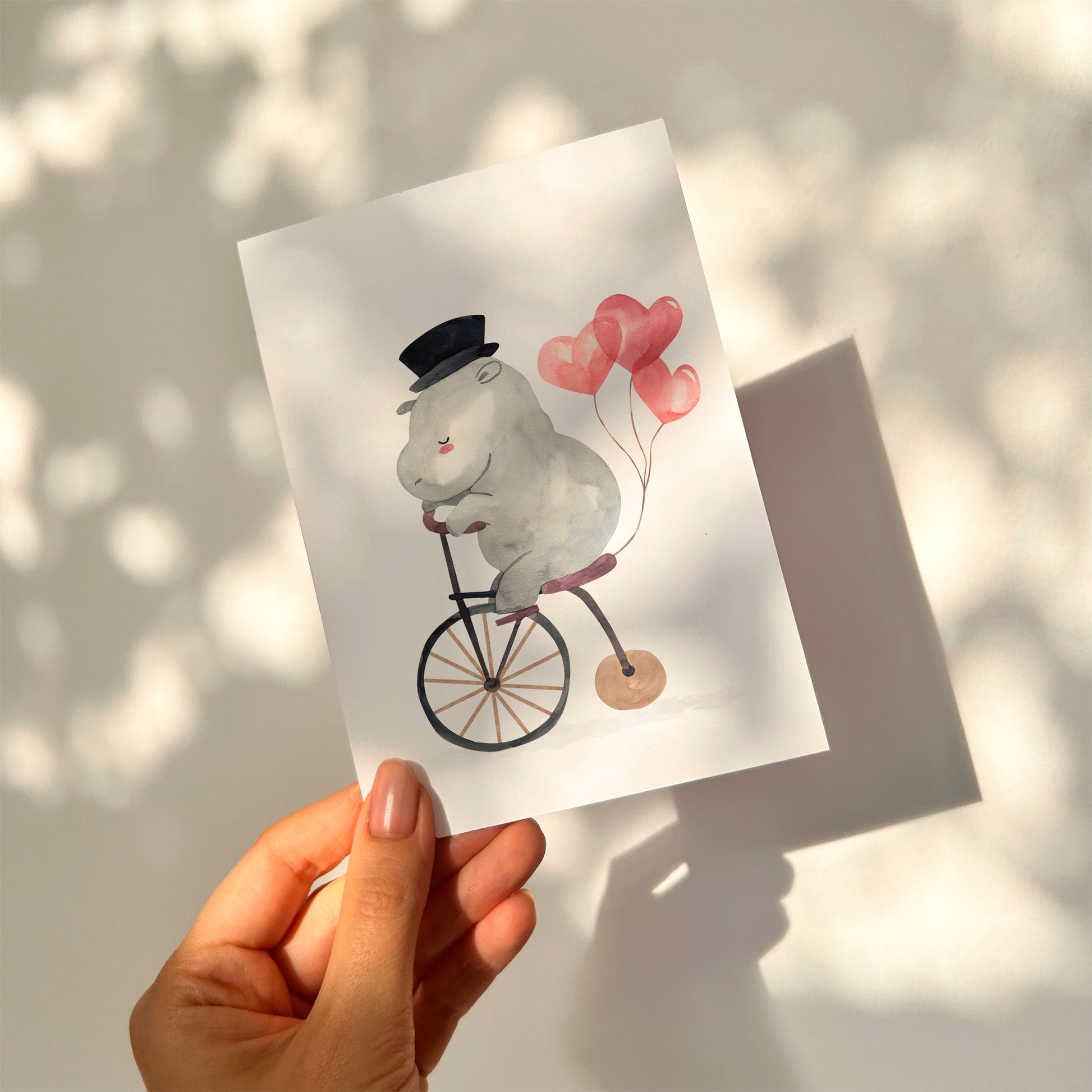 Karte Flusspferd & Fahrrad A6 zeigt ein illustriertes, graues Hippo mit Zylinder auf einem Fahrrad, begleitet von drei Herzballons, gegen eine sonnenbeschienene Wand.