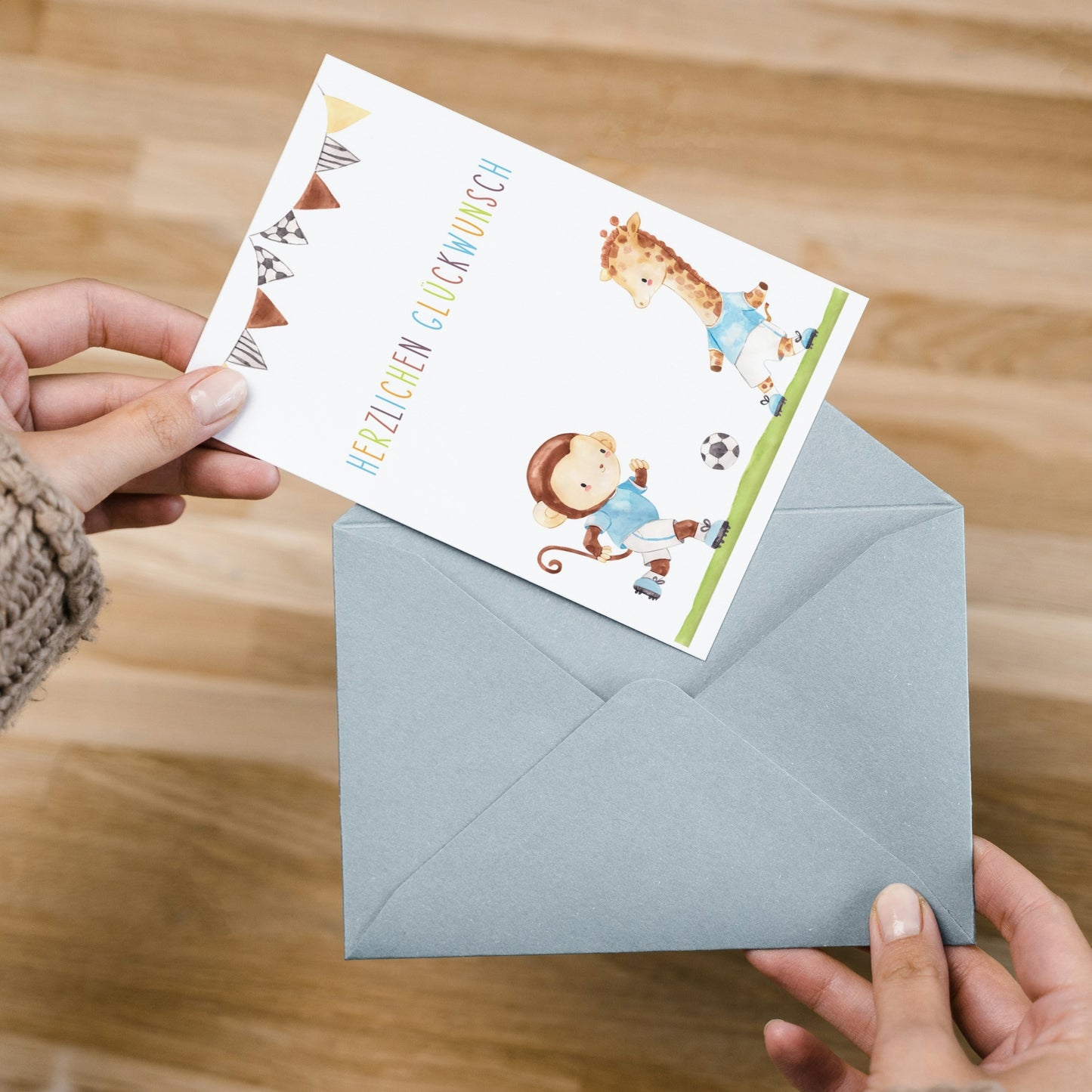 Hände halten Karte Kindergeburtstag Fußball A6 mit halbausgezogener Grußkarte: Aquarell-Pennantflags, Herzlichen Glückwunsch, Cartoon-Affen und Giraffe beim Fußballspielen.