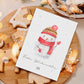Personalisierte Weihnachtskarte Schneemann A6 mit handgemaltem Schneemann, rotem Schal, winterlicher Mütze. Umgeben von Lebkuchenplätzchen und Lichterdekoration auf Holzbrett.