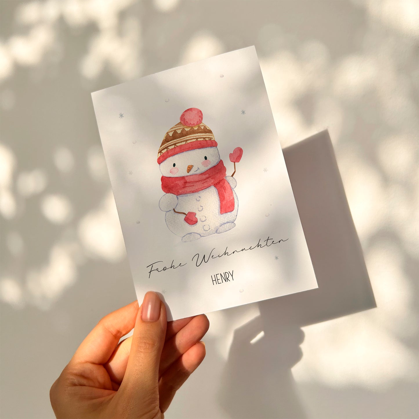 Personalisierte Weihnachtskarte Schneemann A6: Aquarell-Schneemann mit Mütze, rotem Schal und Herz-Fäustlingen, Aufschrift Frohe Weihnachten HENRY, in Hand gehalten.