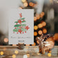 Personalisierte Weihnachtskarte Weihnachtsbaum A6 mit Aquarell-Weihnachtsbaum, Frohe Weihnachten und Namen. Neben goldfarbenem Rentier-Teelichthalter und Tannenzapfen auf Holz.