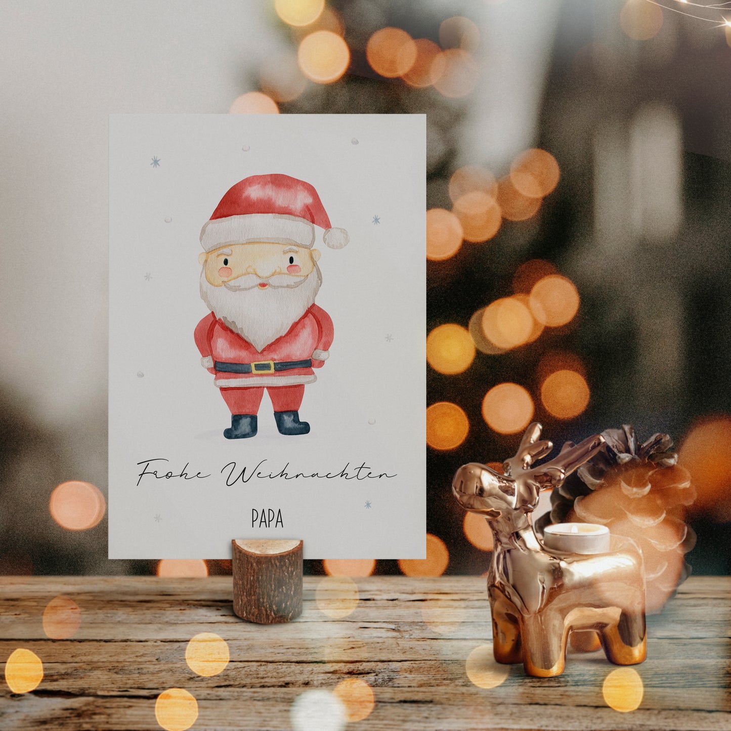 Personalisierte Weihnachtskarte Weihnachtsmann A6 in einem kleinen Holzständer, begleitet von einem goldenen Rentier-Teelichthalter und Tannenzapfen auf rustikalem Holz.