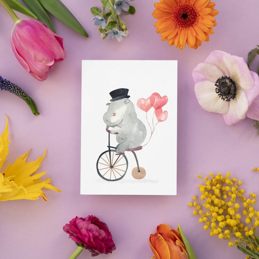 Karte Flusspferd & Fahrrad A6 zeigt ein Hippo auf einem Penny-Farthing mit Herzballons, umgeben von Frühlingsblumen, gedruckt auf hochwertigem Naturpapier.