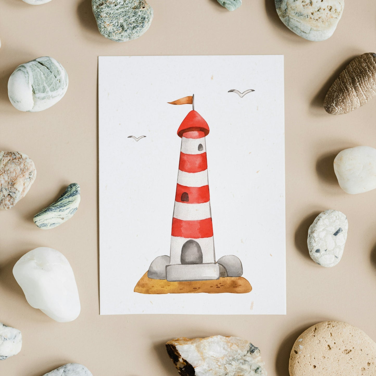 Postkarte Leuchtturm A6 auf beigem Untergrund mit Steinen, zeigt einen illustrierten Leuchtturm auf einer Insel, ideal für maritime Grüße oder Dekoration.