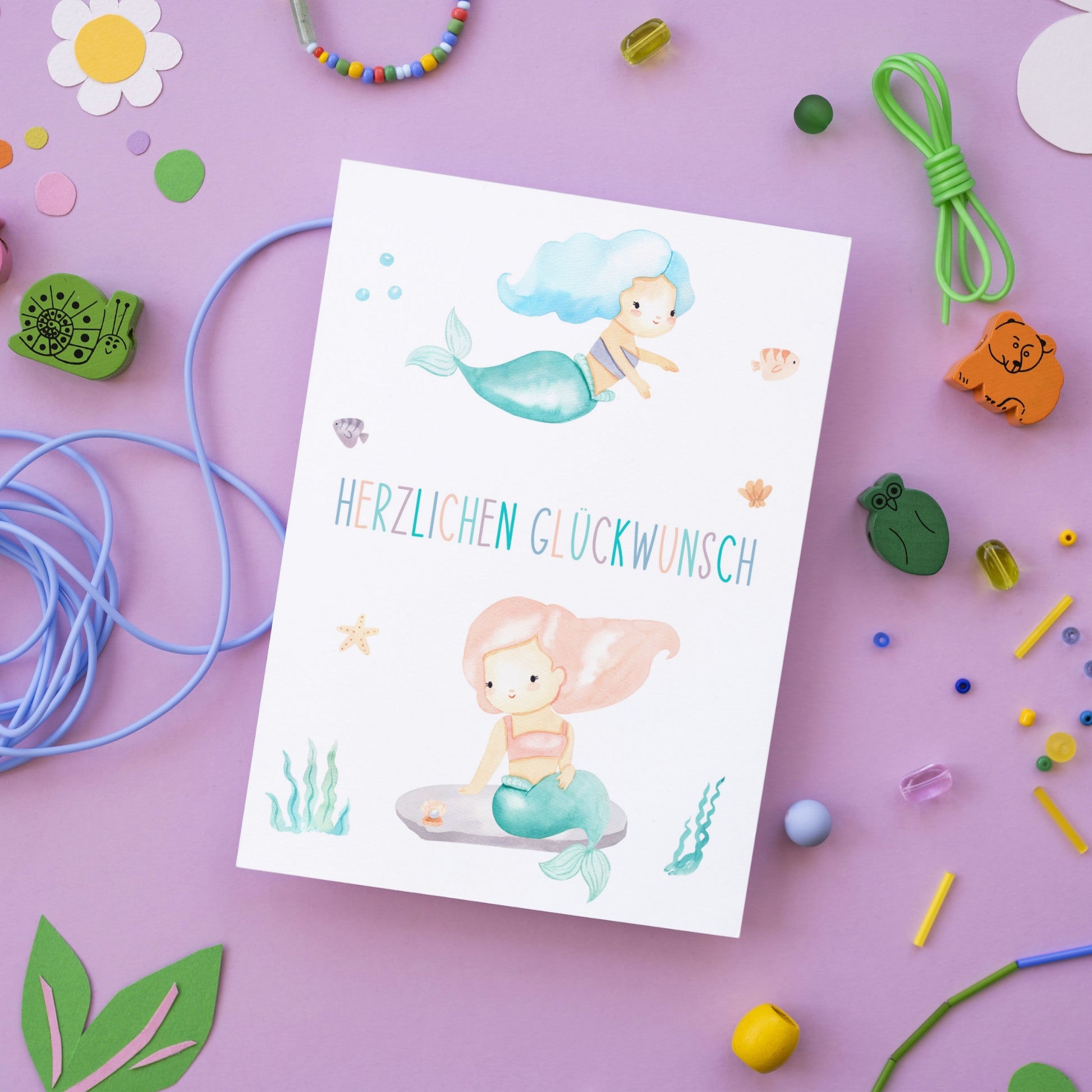 Karte Kindergeburtstag Meerjungfrauen mit handgemalten Meerjungfrauen, Herzlichen Glückwunsch Schriftzug, dekoriert mit Seestern, Seegras und Fischen, ideal für kleine Meerjungfrauen-Fans.