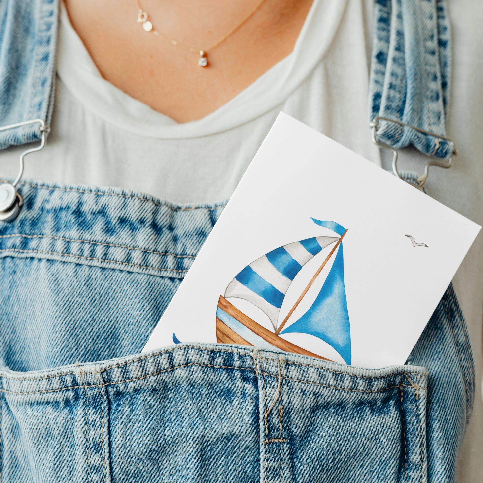 Nahaufnahme von jemandem in hellen Jeansoveralls, aus deren Latz die Postkarte Postkarte Segelboot A6 mit einem Segelboot-Aquarell herausragt.