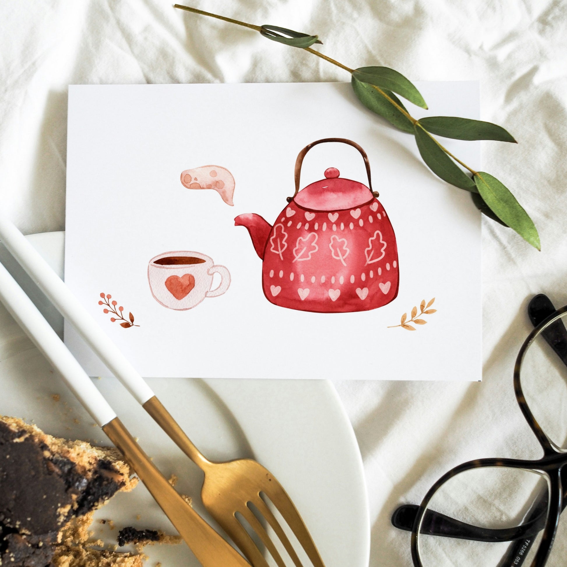 Die Postkarte Karte Tea Time A6 zeigt eine Aquarell-Teekanne und Tasse mit Herzmotiven, umgeben von dekorativen Zweigen und liegt auf weißem Stoff.