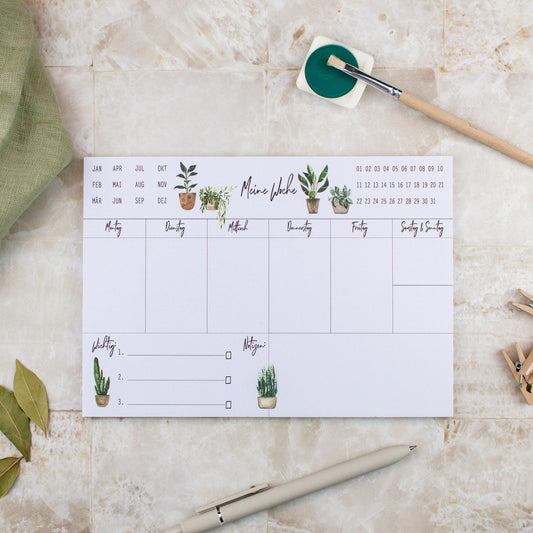 Wochenplaner Pflanzen A5 Block: Ein Wochenplaner mit handgemalten Pflanzenillustrationen, einem Stift und Pinsel auf Papier, perfekt für Termine, Notizen und To-dos.