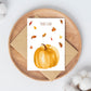 Postkarte Karte Kürbis & Herbstlaub Herbstliebe A6 auf einem Teller, umgeben von herbstlichen Blättern und einem Kürbis, auf hochwertigem Naturpapier gedruckt.