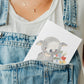 Person in Jeanslatzhose hält eine Karte, „Karte Blumengrüße Koala A6“, mit Koala-Motiv, gedruckt auf mattem 300g Naturpapier, in der Tasche.