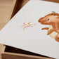 Poster Feuerdrache A3 & A4: Deckel einer Holzkiste mit einem weißen Blatt, darauf ein niedlicher, aquarellierter Cartoon-Dinosaurier, der stilisierte Flammen ausstößt.