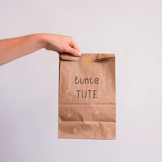 Hand hält eine braune Papiertüte mit schwarzem Text, die 10 A6-Karten mit leichten Fehlern enthält. Produkt: Bunte Tüte mit 10 Karten A6.