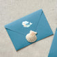 Sticker Meerjungfrau blau auf blauem Briefumschlag mit Muschel, zeigt handgemalten Meerjungfrau-Sticker in Pastelltönen. Ideal zur Dekoration von Notizbüchern und Laptops.