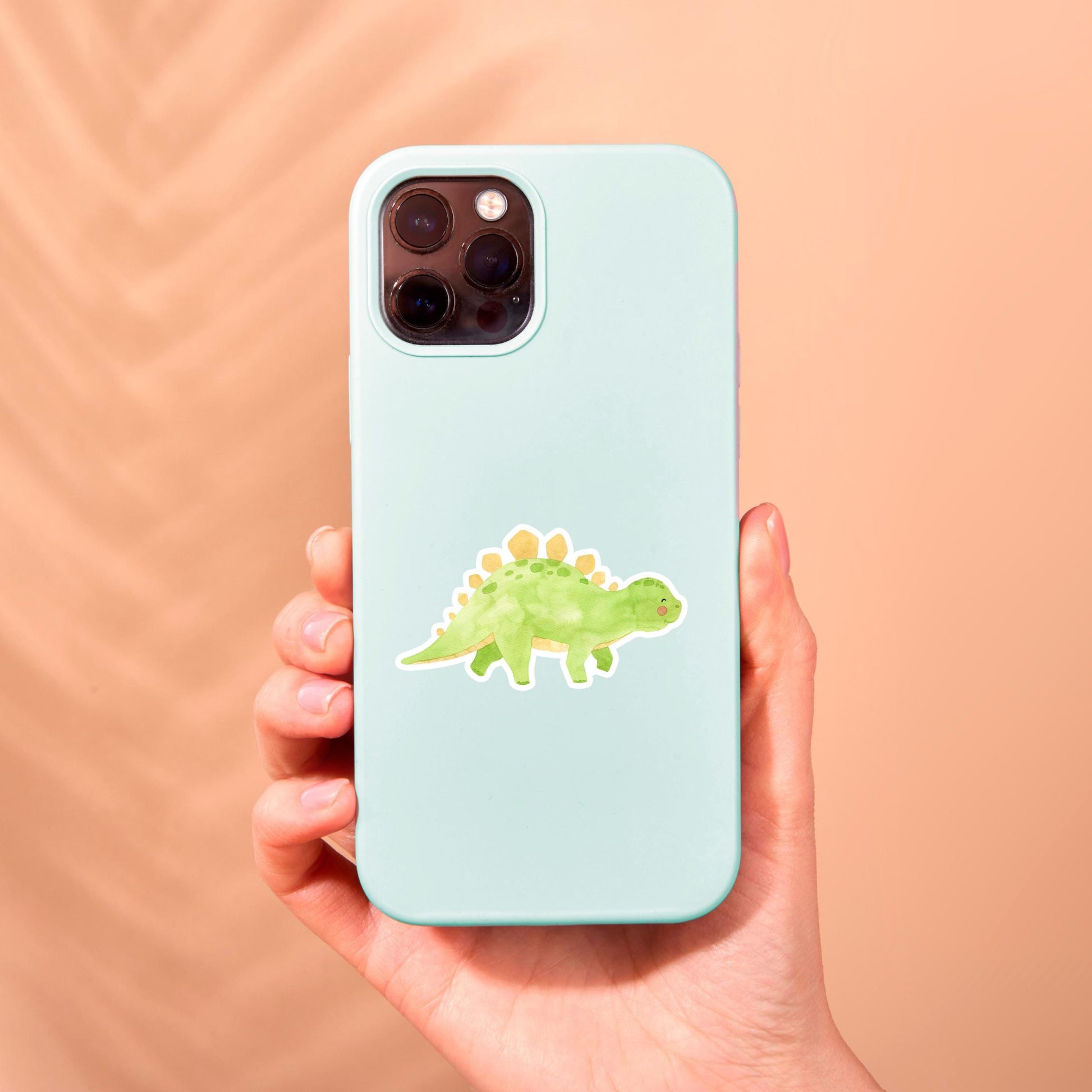 Hand hält Handyhülle mit Sticker Dinosaurier Stego, einem handgefertigten, spritzfesten Vinyl-Aufkleber in Form eines grünen Dinosauriers mit gelben Flecken.