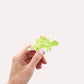 Sticker Dinosaurier Stego: Hand hält einen handgemalten Vinyl-Dino-Sticker. Ideal für Bullet Journals, Laptops oder Bastelprojekte. Spritzfest und langlebig.