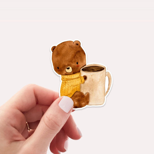Hand hält Sticker Bär & Kaffee: Ein Vinyl-Aufkleber mit handgemaltem Bären, der eine Tasse hält, ideal für kreative Dekorationen.