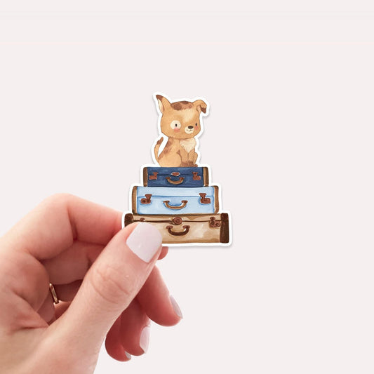 Hand hält Sticker Hund & Koffer, zeigt handgemalten Cartoon-Hund auf Gepäckstapel. Vinyl, spritzfest, ideal für Bullet Journals, Laptops, Handyhüllen.
