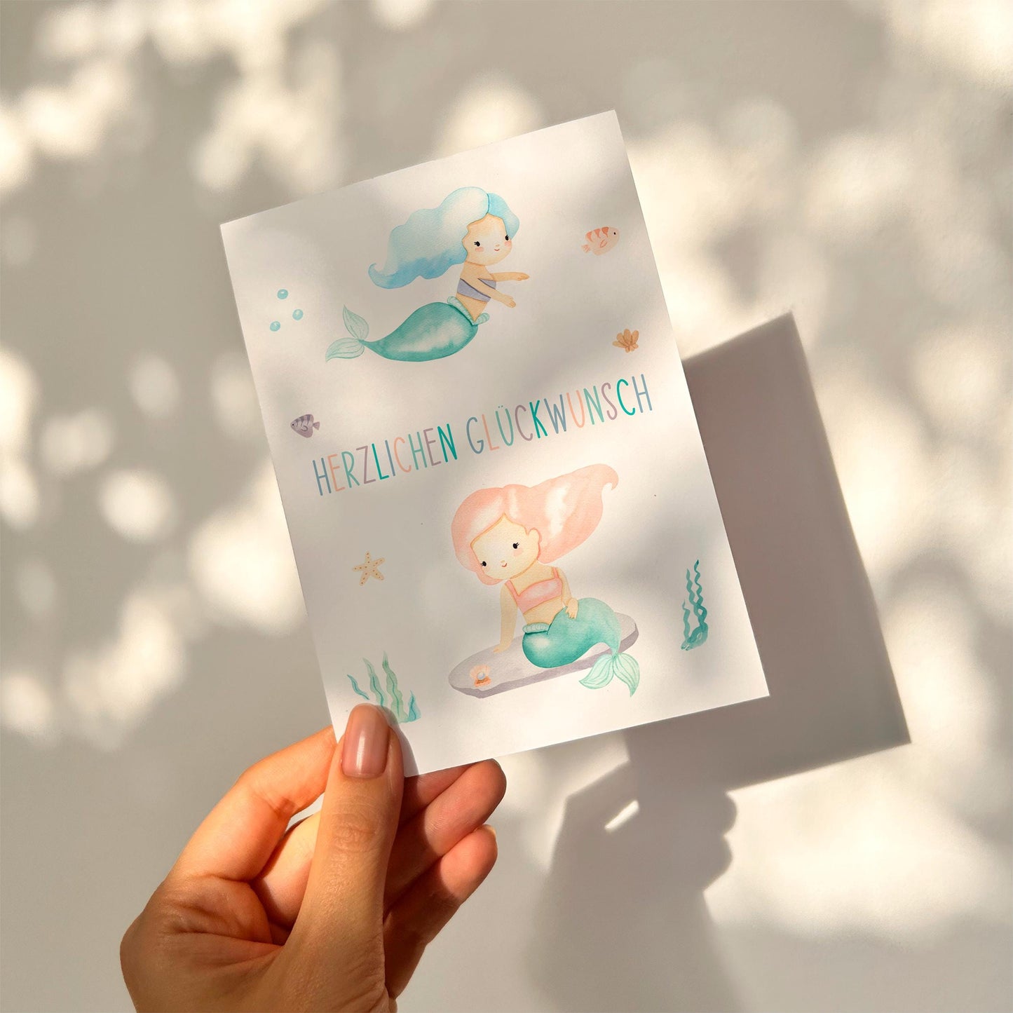 Hand hält Geburtstagskarte Kindergeburtstag Meerjungfrauen mit handgemalten Meerjungfrauen und 'Herzlichen Glückwunsch' Schriftzug auf hochwertigem Naturpapier. Ideal für kleine Meerjungfrauen-Fans.