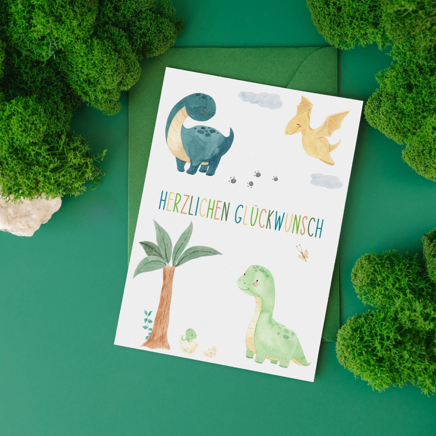 Karte Kindergeburtstag Dinosaurier A6 mit handgemalten Dinosauriern und Herzlichen Glückwunsch Schriftzug, inklusive grünem Umschlag auf grünem Hintergrund.