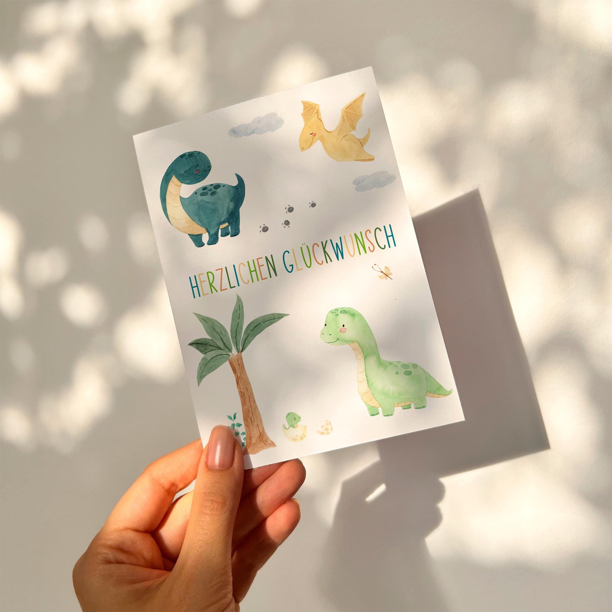 Karte Kindergeburtstag Dinosaurier A6 mit handgemalten Dinos und Herzlichen Glückwunsch Schriftzug, gehalten von einer Hand, auf stabilem Naturpapier gedruckt.
