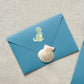 Sticker Dinosaurier Tyranno: Blauer Umschlag mit Dinosaurier- und Muschel-Sticker. Handgezeichnet, spritzfest, ideal für Bullet Journals oder Einladungen. Größe: ca. 6 x 4,5 cm.