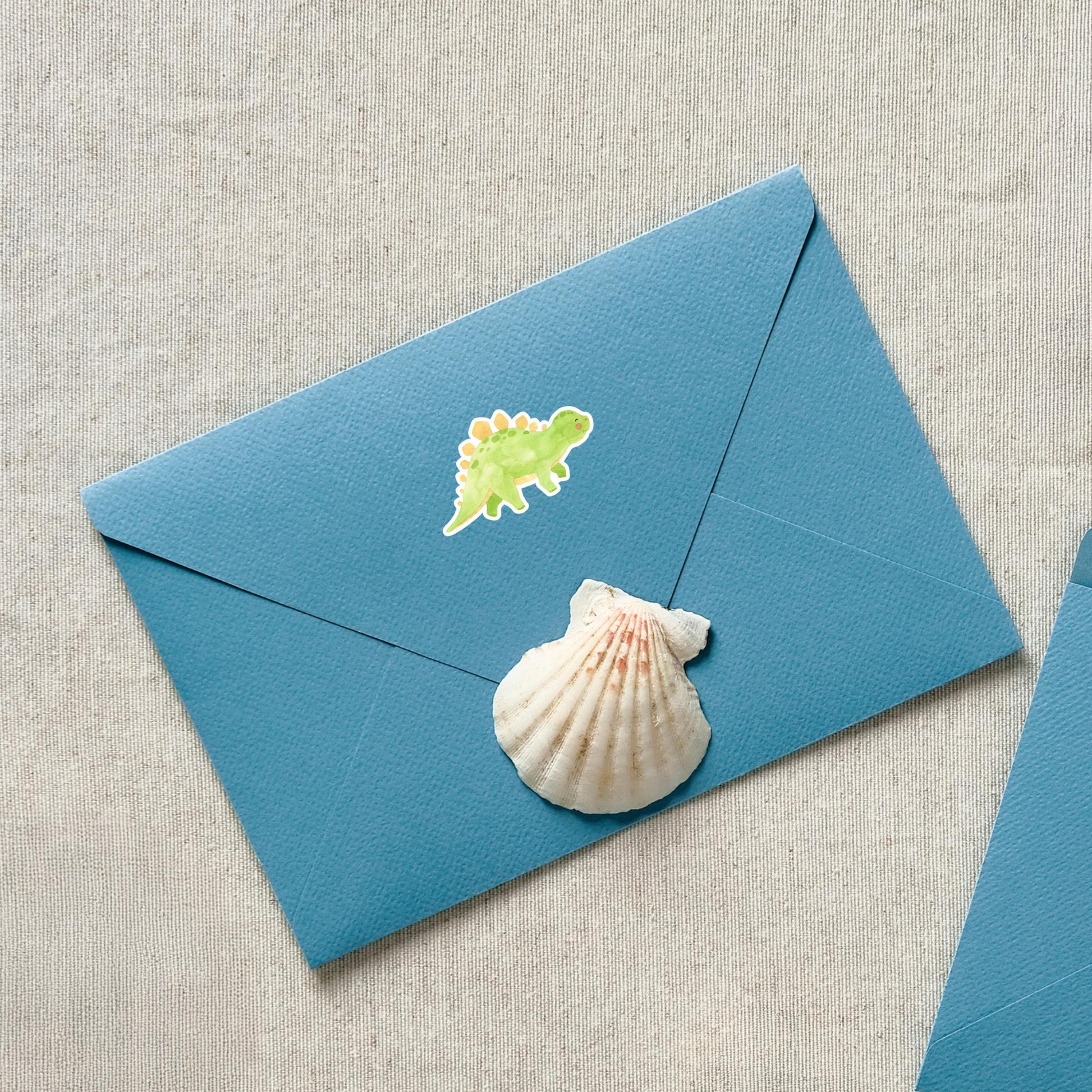 Sticker Dinosaurier Stego auf einem blauen Umschlag mit Muschel-Aufkleber. Handgemalter, spritzfester Vinyl-Sticker perfekt für Bullet Journals oder Einladungen.