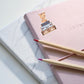 Sticker Hund & Koffer: Handgemalter Hunde-Sticker auf einem rosa Notizbuch mit Bleistiften, inspiriert zu Reisen, ideal für Bullet Journals und DIY-Projekte.