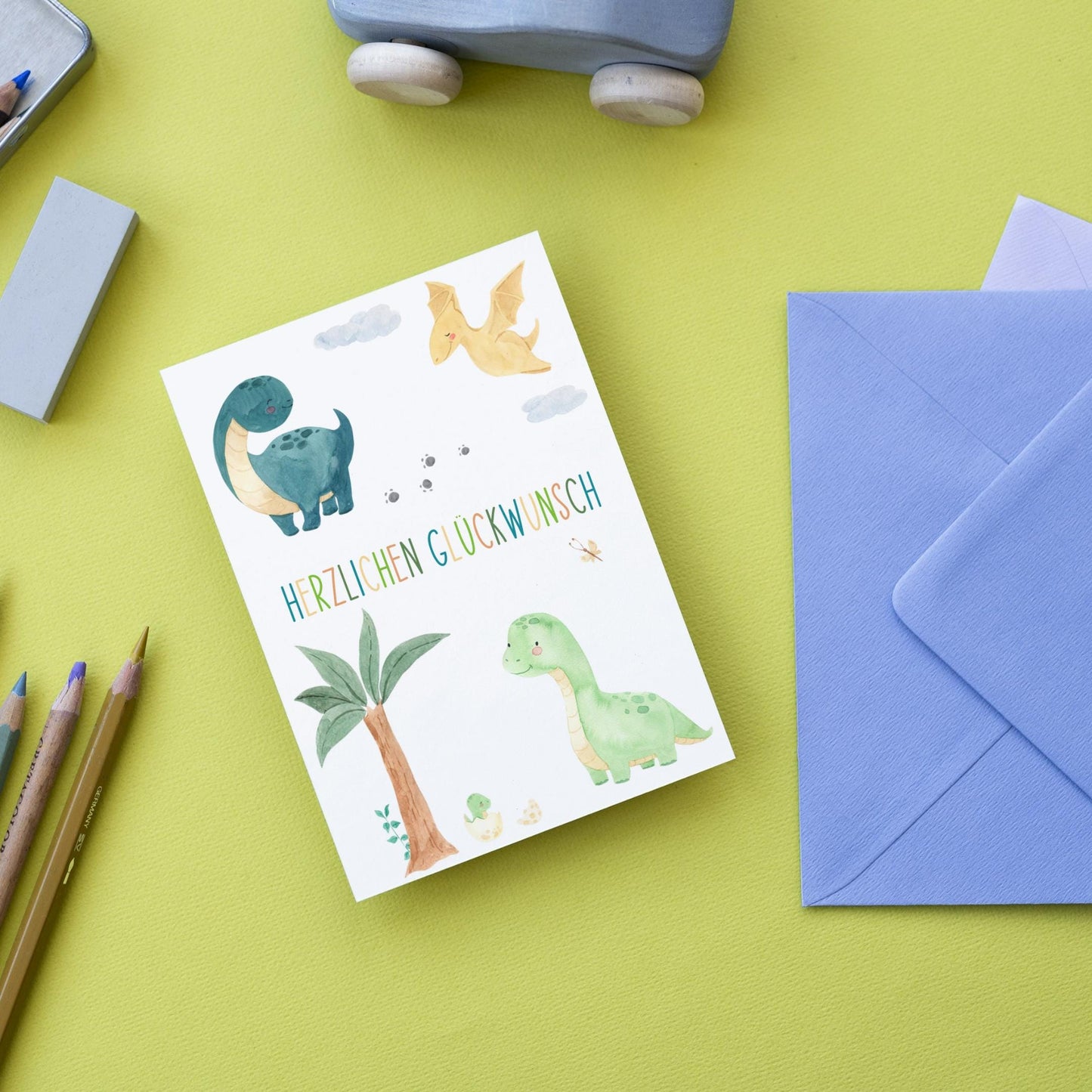 Karte Kindergeburtstag Dinosaurier A6 mit handgemalten Dino-Motiven und Schriftzug, begleitet von einem blauen Umschlag und Stiften, perfekt für kleine Dino-Fans.