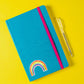 Geschlossener Notizbuch mit Sticker Regenbogen auf dem Cover, daneben ein Kugelschreiber. Der handgemalte Vinyl-Sticker ist spritzfest und langlebig.