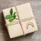 Geschenk in Kraftpapier mit Juteschnur gebunden, verziert mit einem frischen Zweig und einem Sticker Regenbogen, handgemalt, wasserfest, ideal für kreative Dekorationen.
