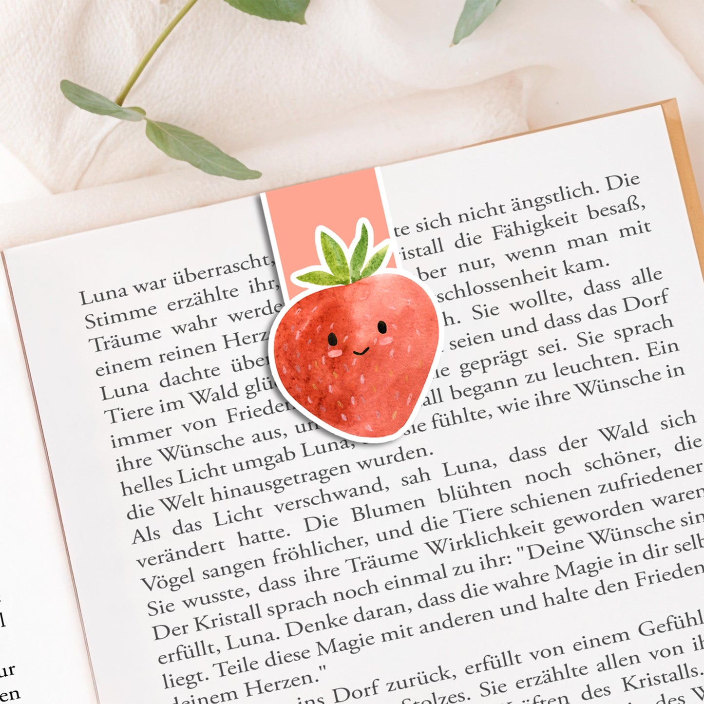 Magnetisches Lesezeichen Erdbeere auf offenem Buch, handgemalt, auf einer Stoffoberfläche mit Pflanzenzweigen platziert, zeigt einen versiegelten, langlebigen Buchmarker.