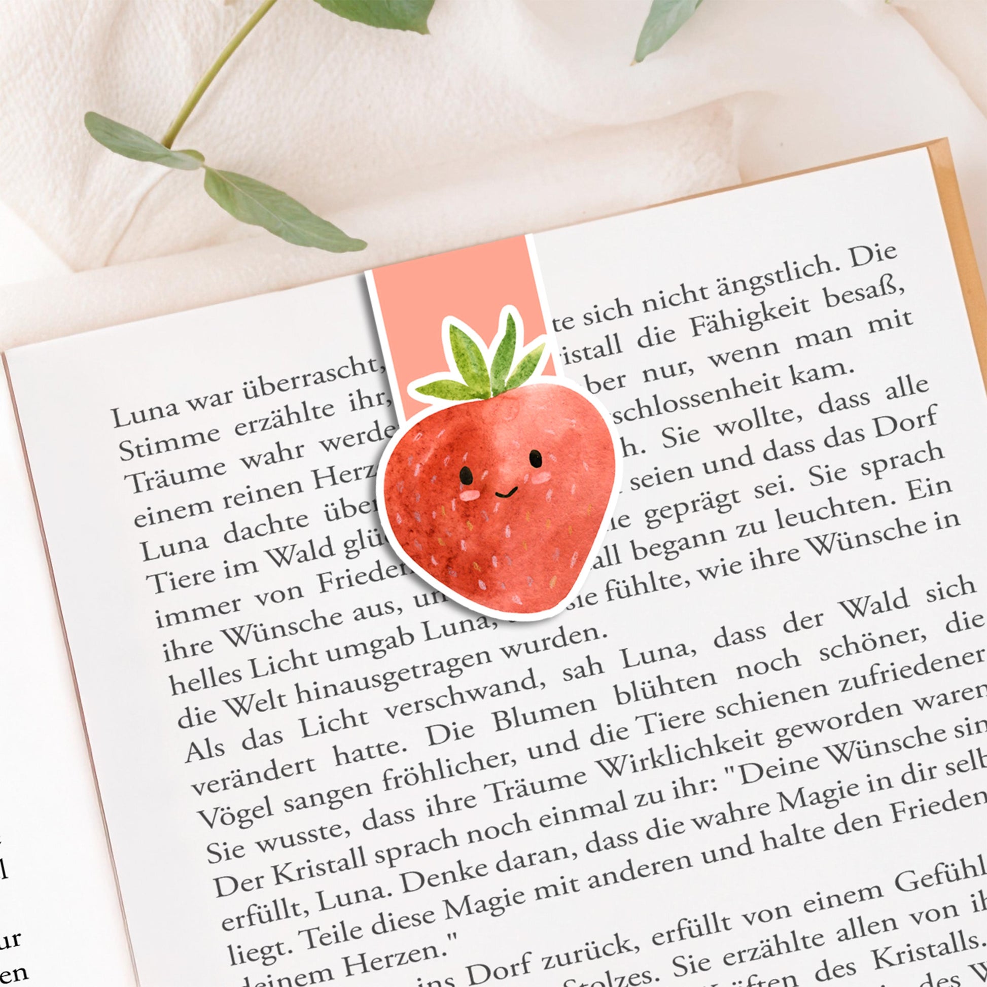 Magnetisches Lesezeichen Erdbeere auf offenem Buch, handgemalt, auf einer Stoffoberfläche mit Pflanzenzweigen platziert, zeigt einen versiegelten, langlebigen Buchmarker.