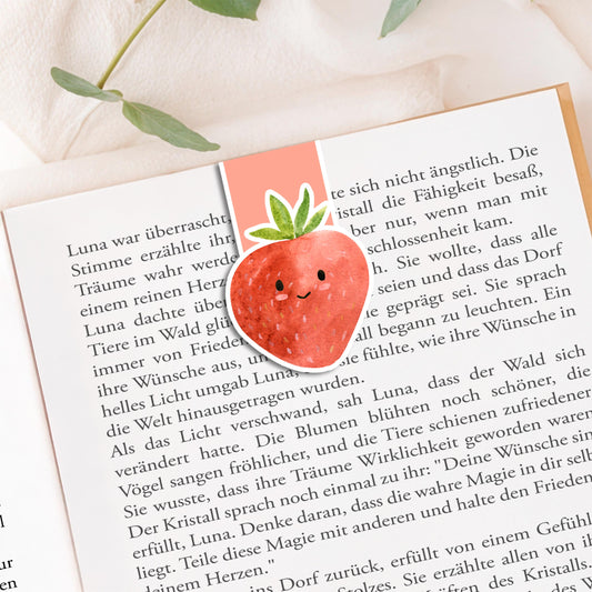 Magnetisches Lesezeichen Erdbeere auf offenem Buch, handgemalt, auf einer Stoffoberfläche mit Pflanzenzweigen platziert, zeigt einen versiegelten, langlebigen Buchmarker.