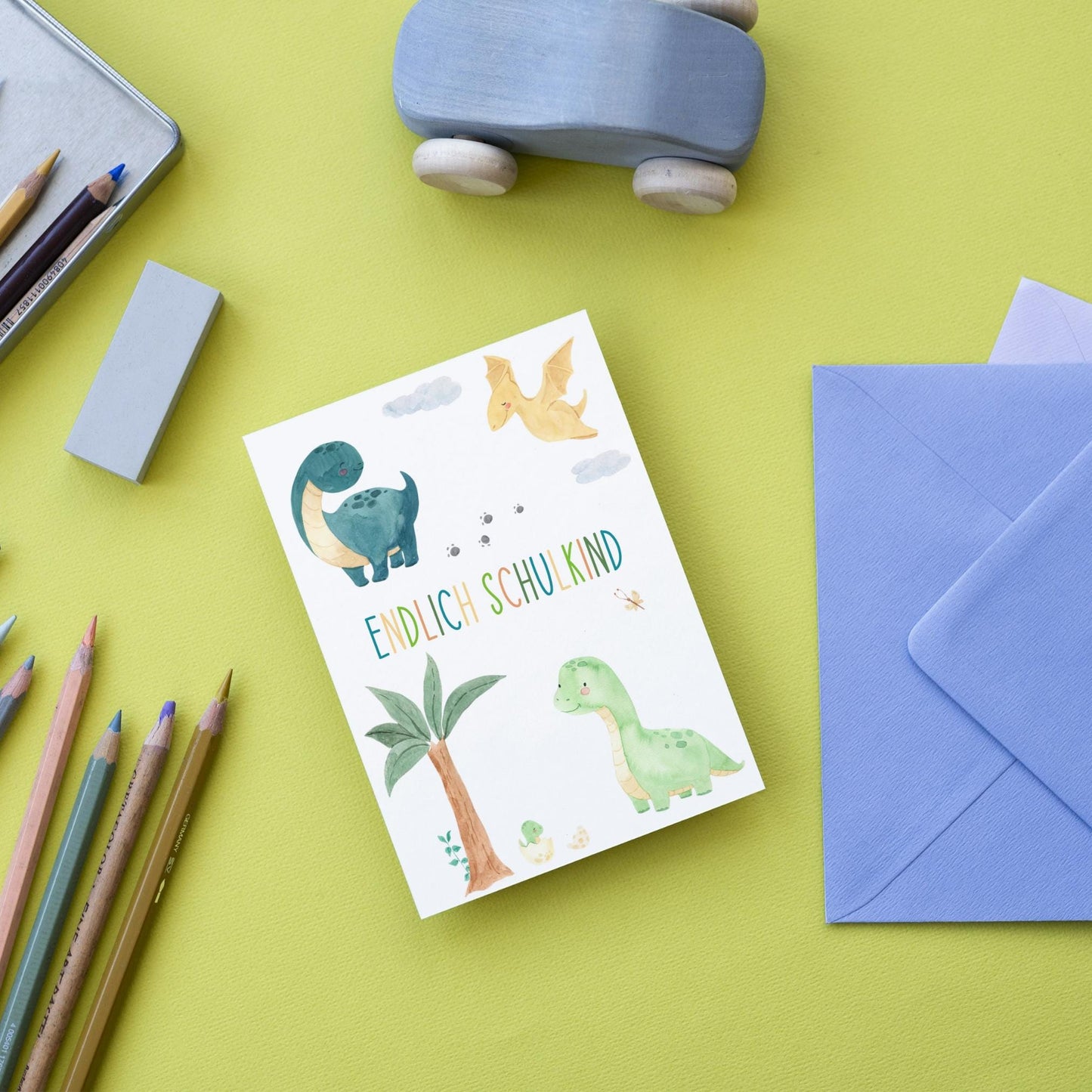 Karte Endlich Schulkind Dinosaurier A6 mit Aquarell-Dinosauriern, ideal zur Einschulung. Umgeben von Buntstiften, Radiergummi, Spielzeugauto und blauem Umschlag.