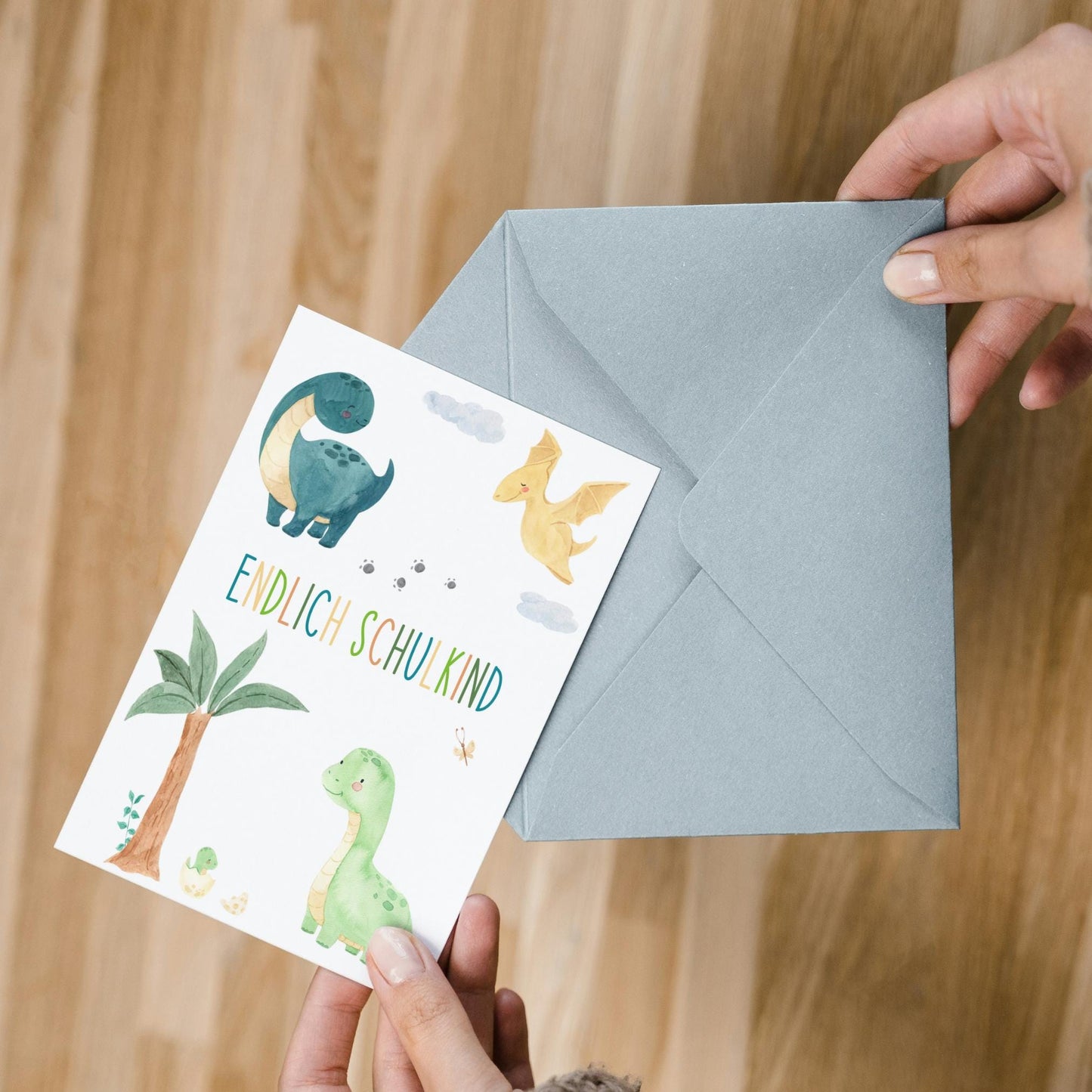 Karte Endlich Schulkind Dinosaurier A6, zeigt süße Aquarell-Dinos auf weißem Hintergrund, wird in oder aus einem hellblauen Umschlag geschoben. Ideal zur Einschulung.