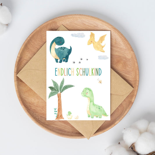 Karte Endlich Schulkind Dinosaurier A6 auf Holztablett, mit Dino-Motiven und Endlich Schulkind-Text, inkl. Umschlag, umgeben von Baumwollzweigen als Dekoration.