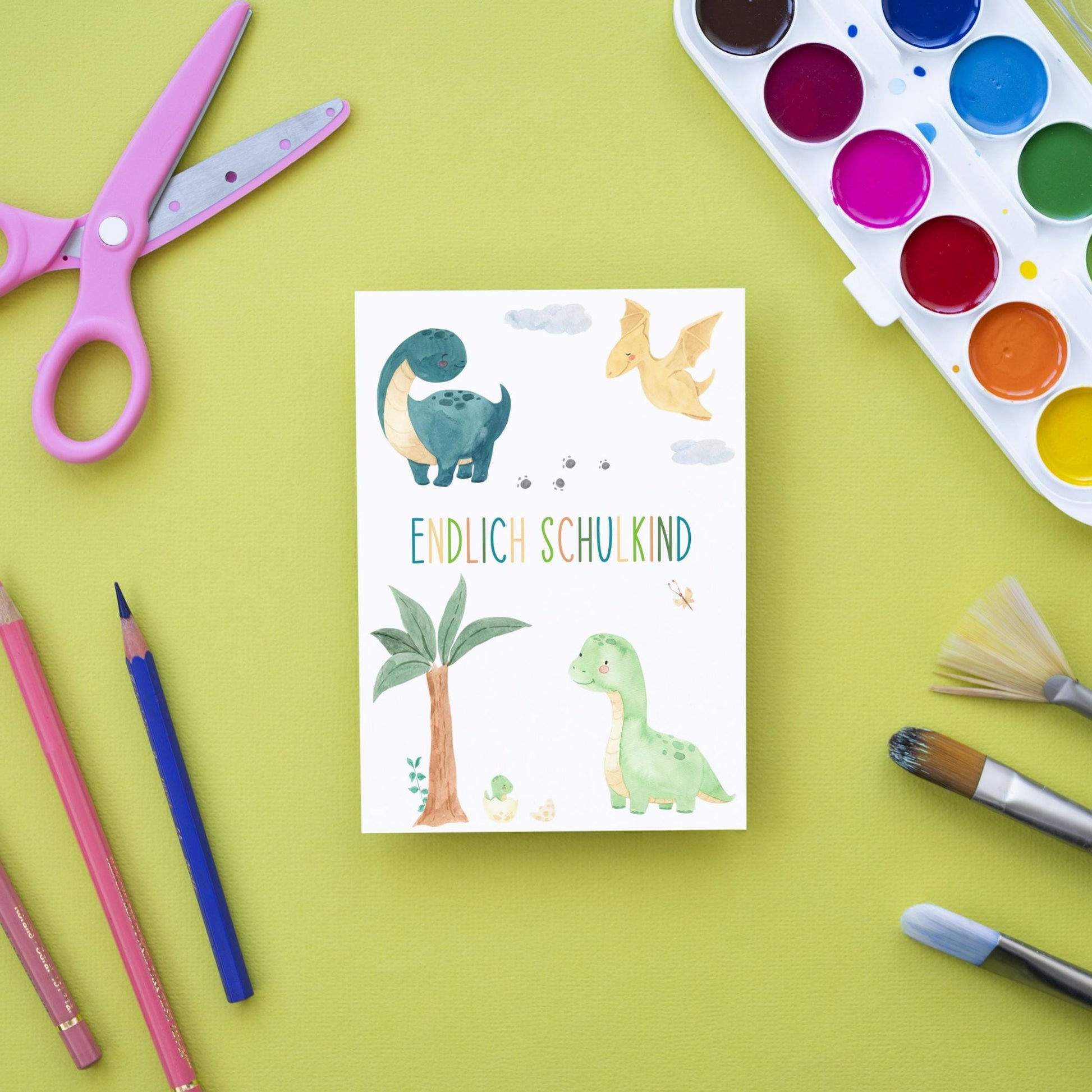 Karte Endlich Schulkind Dinosaurier A6 auf grünem Untergrund, umgeben von Bastelutensilien. Vier Cartoon-Dinos auf A6-Karte, ideal zur Einschulung.